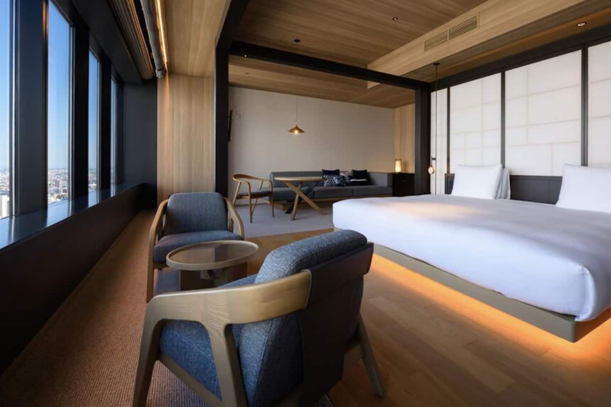 Une chambre d’hôtel moderne dotée d’un grand lit, d’un coin salon avec des fauteuils, d’une petite table et offrant une vue sur la ville grâce à de grandes fenêtres.