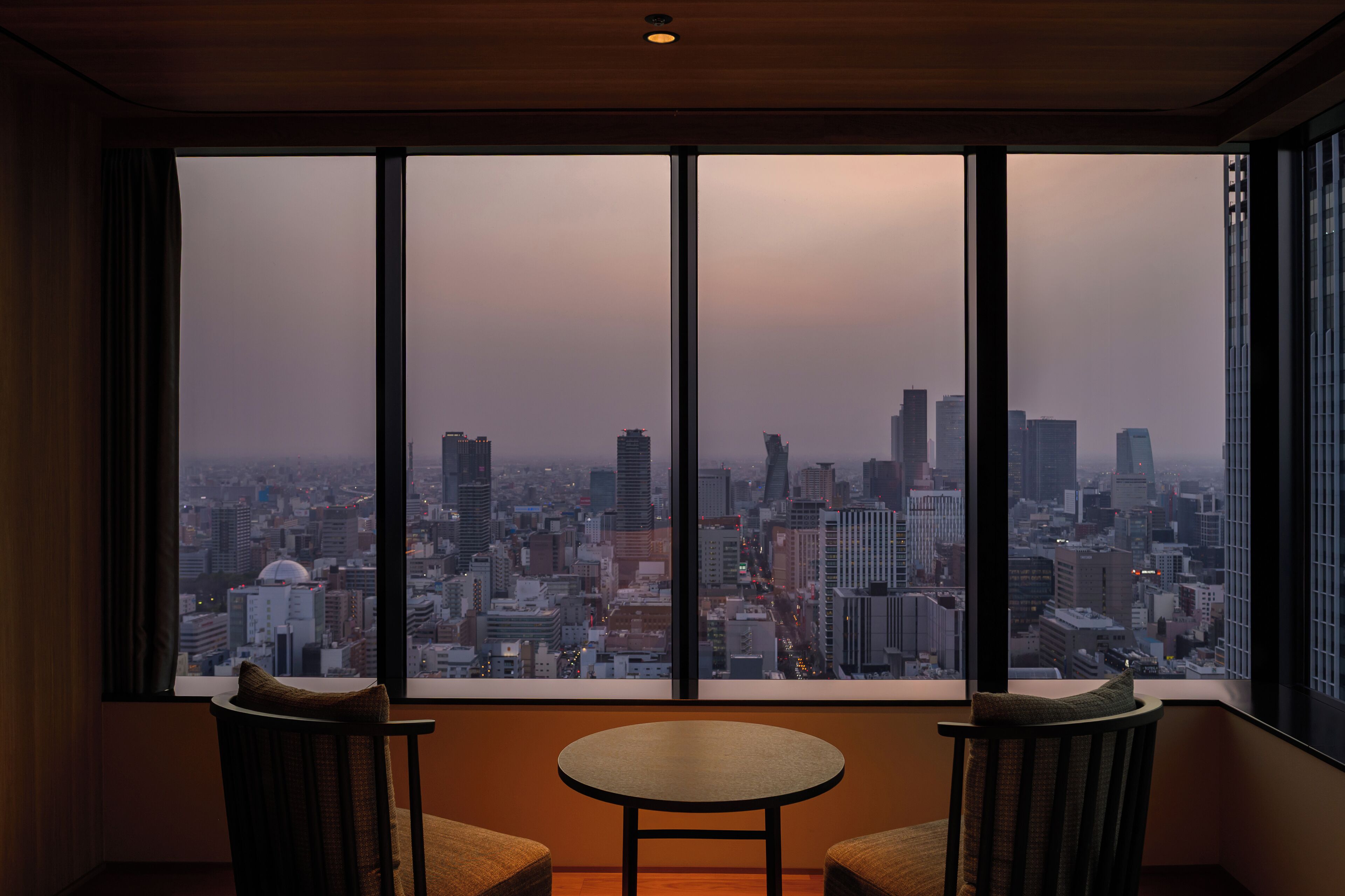 A room with a large window offering a cityscape view. There are two chairs and a small table in the room.