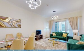 A spacious living room with a green sofa, a dining area with chairs, a TV, and a painting on the wall. There are curtains and a lamp.