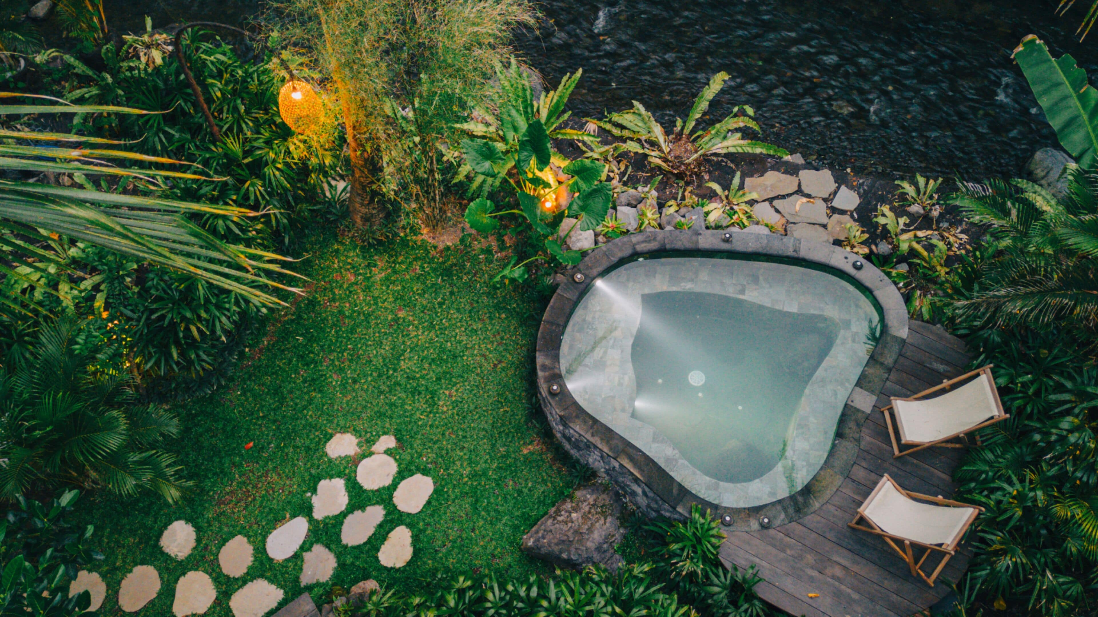 Un bain à remous entouré d’une terrasse en pierre, avec quelques chaises et un petit coin jardin.