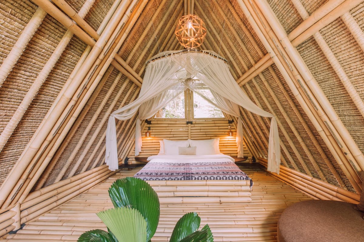 Une pièce dotée d’un plafond en bambou incliné et très haut, d’un lit avec une literie blanche et d’un luminaire suspendu.