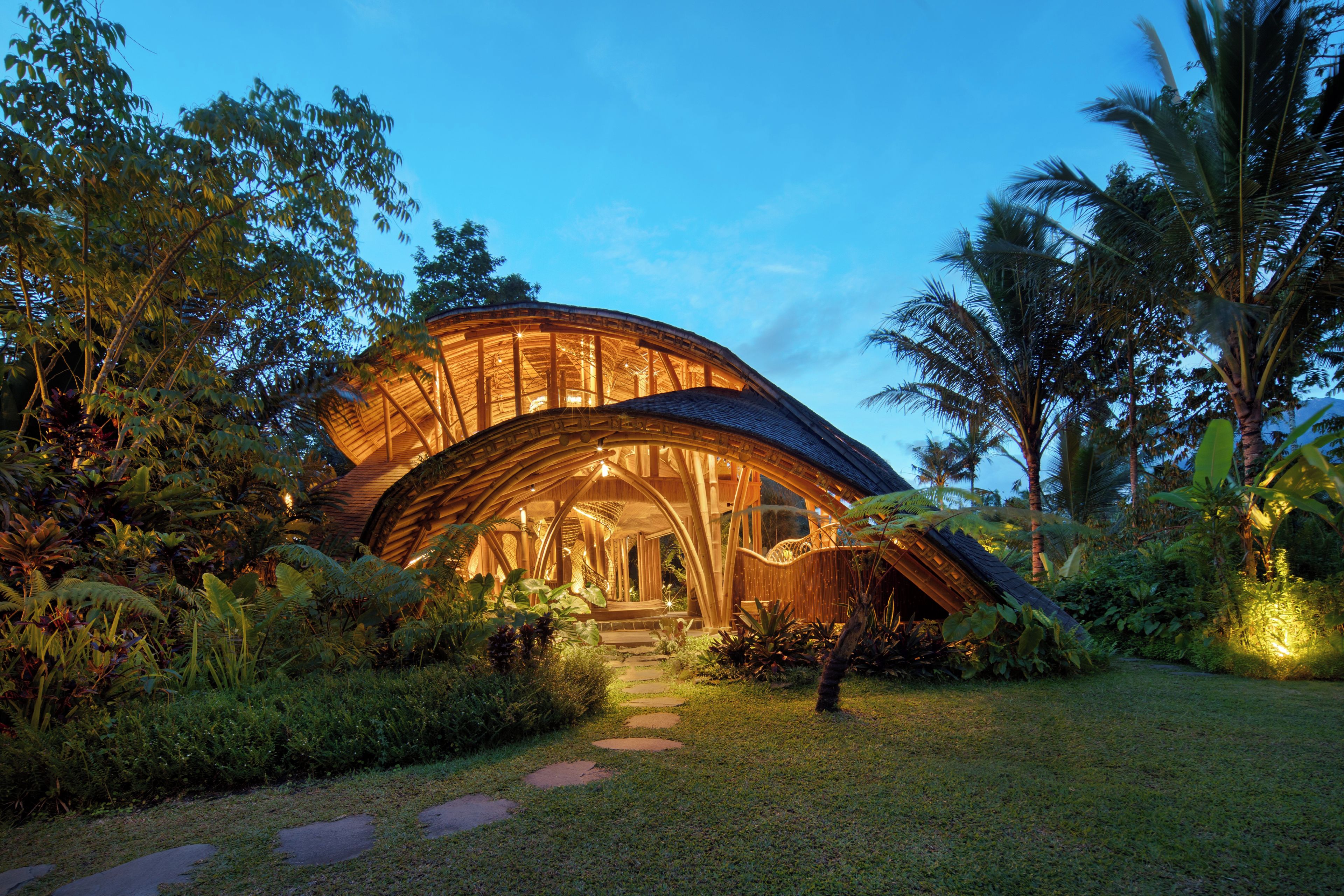 A large, curved building with a wooden exterior and a glass facade, surrounded by lush greenery and palm trees.