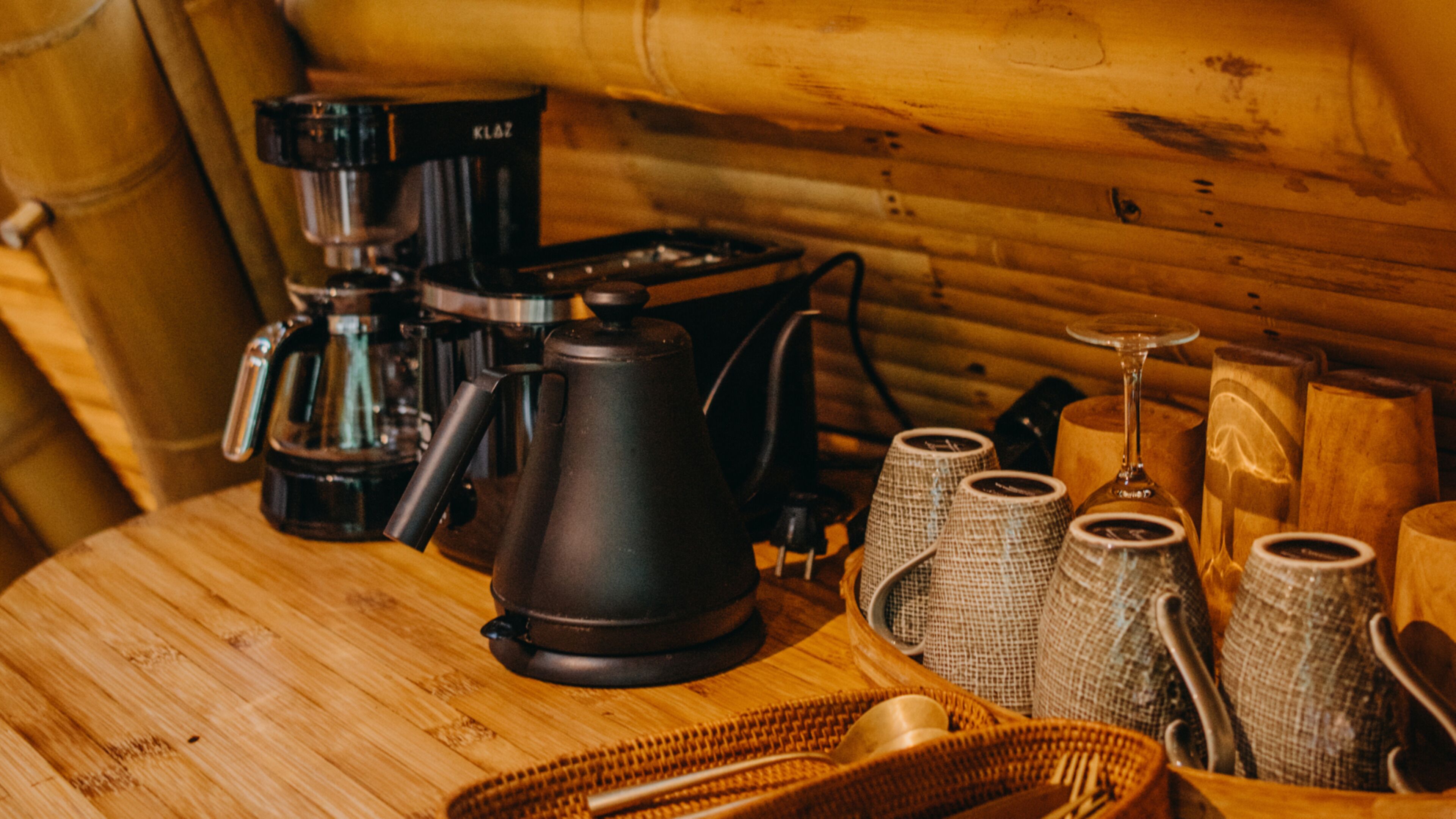 A wooden table with a coffee maker, a kettle, and several cups and glasses.