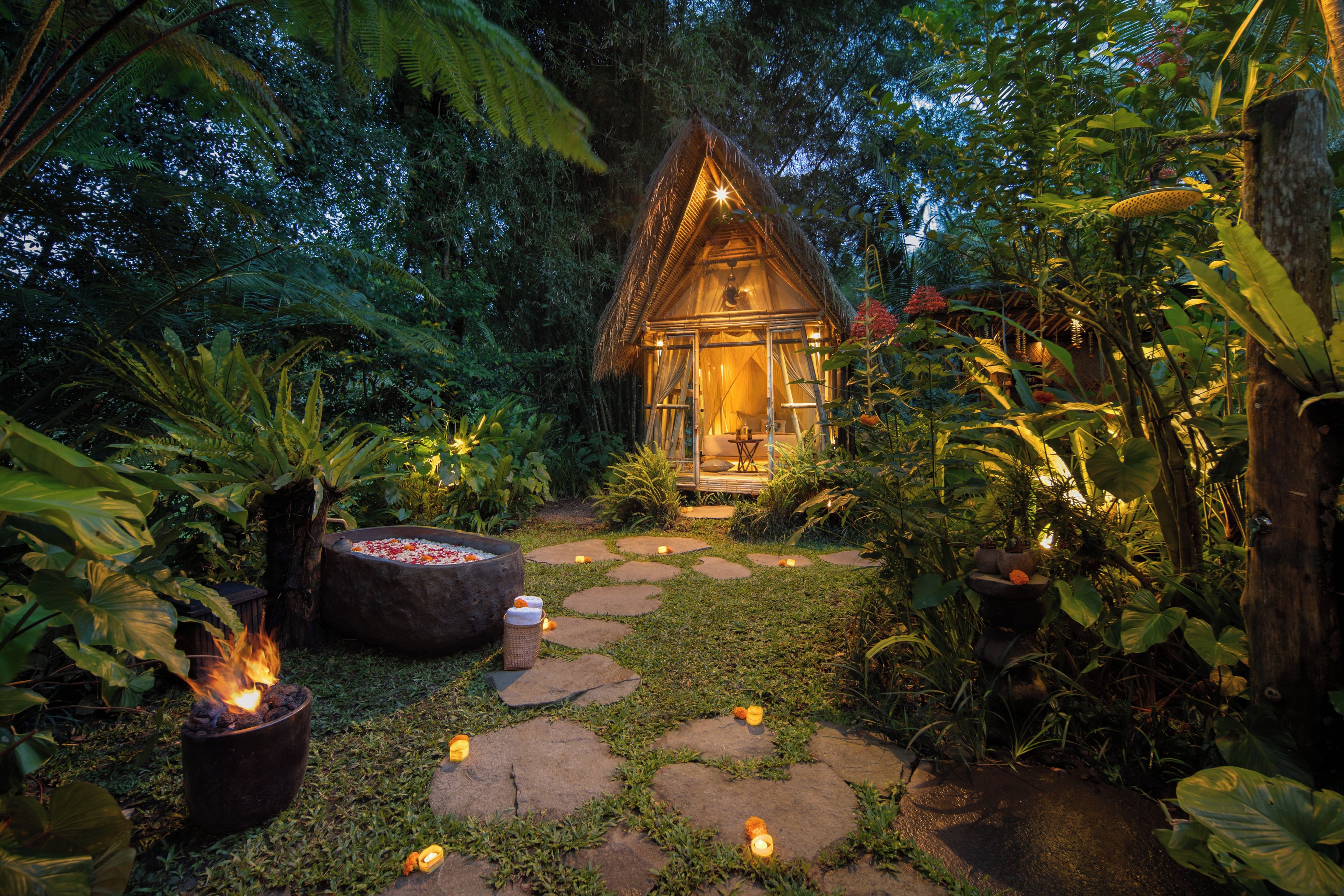 A thatched-roof hut with a fire pit and candles in a lush garden.