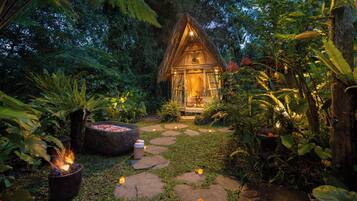 A thatched-roof hut with a fire pit and candles in a lush garden.
