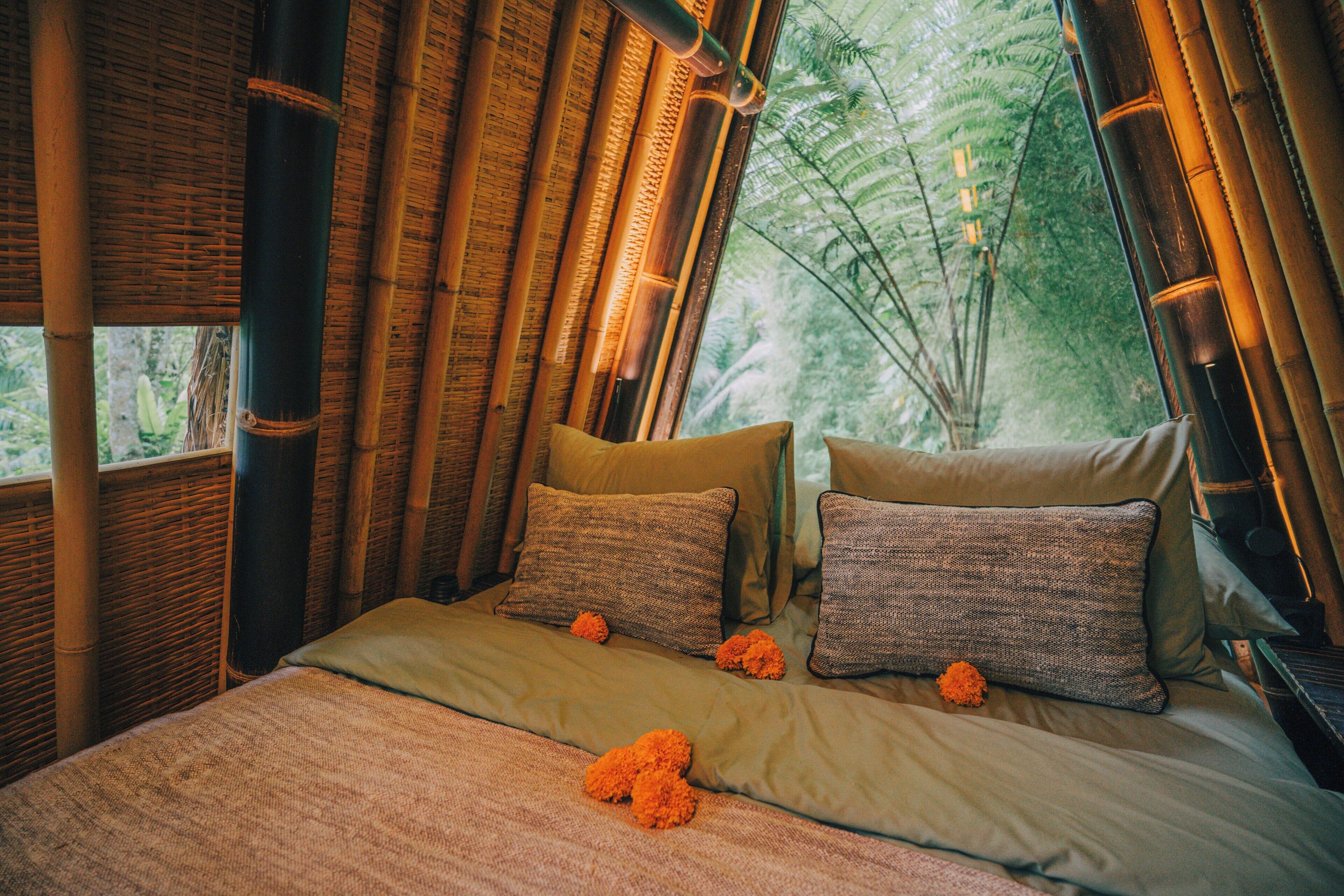 A bed with a green bedspread and two pillows, a window with bamboo blinds, and a view of trees outside.