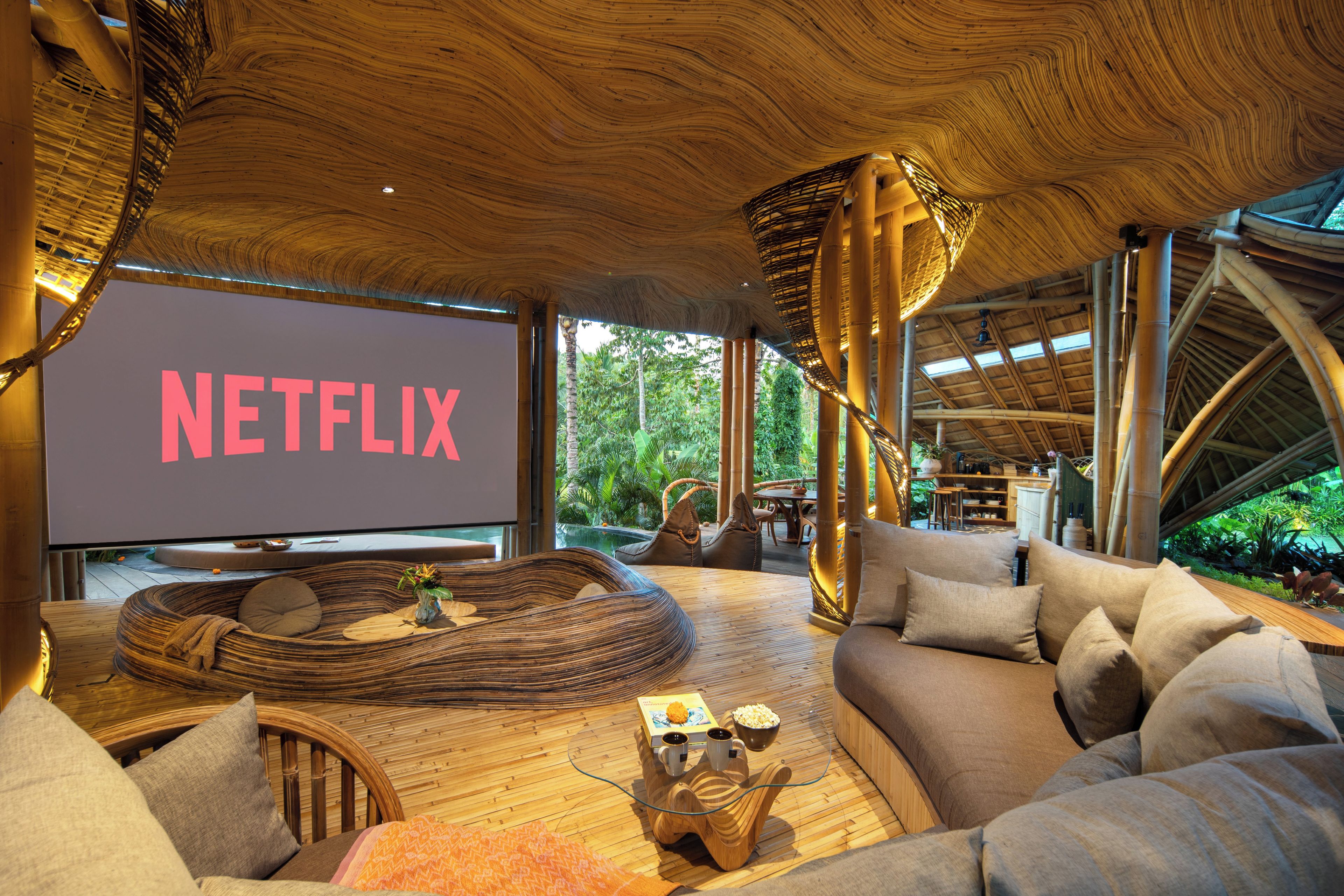 A cozy lounge area with a large screen displaying 'NETFLIX,' comfortable seating, and a view of lush greenery outside.
