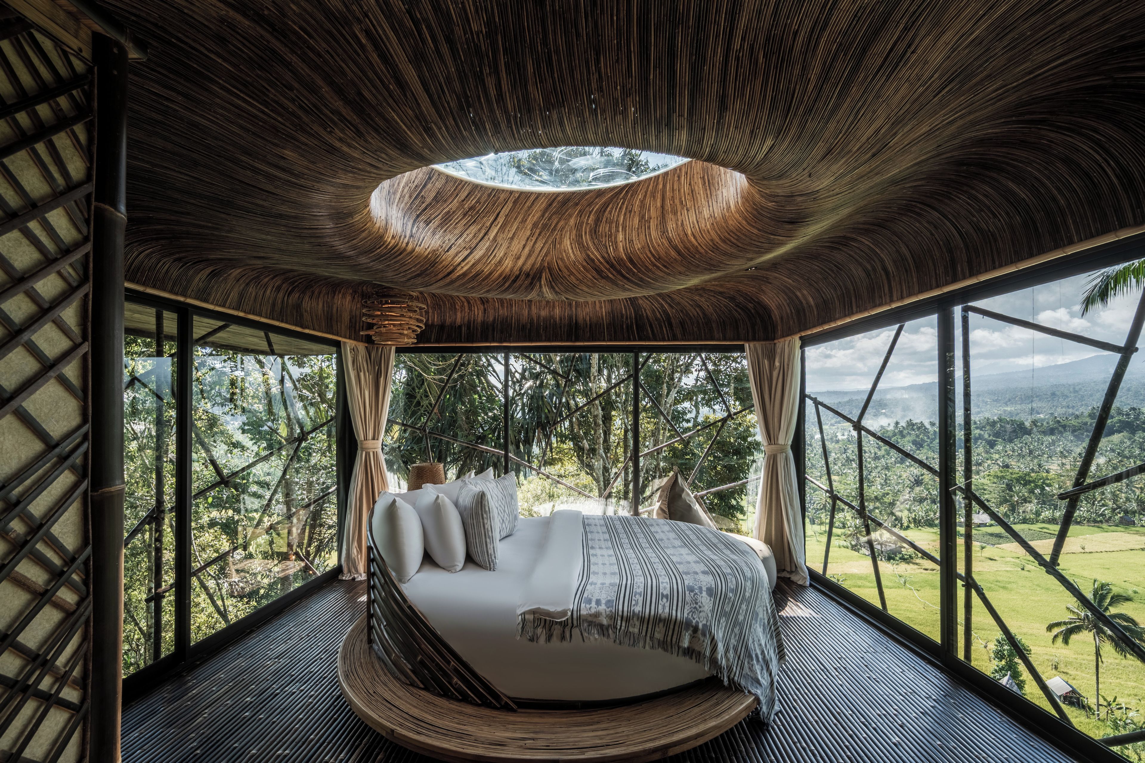 A bed in a room with a view of a forest and a unique ceiling design.