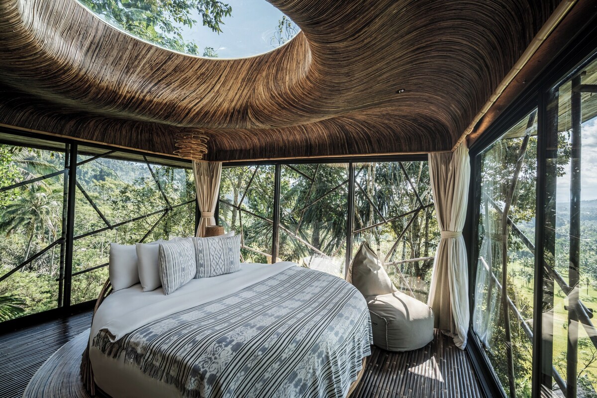 A modern bedroom with a large bed, a view of the forest, and a unique curved ceiling.