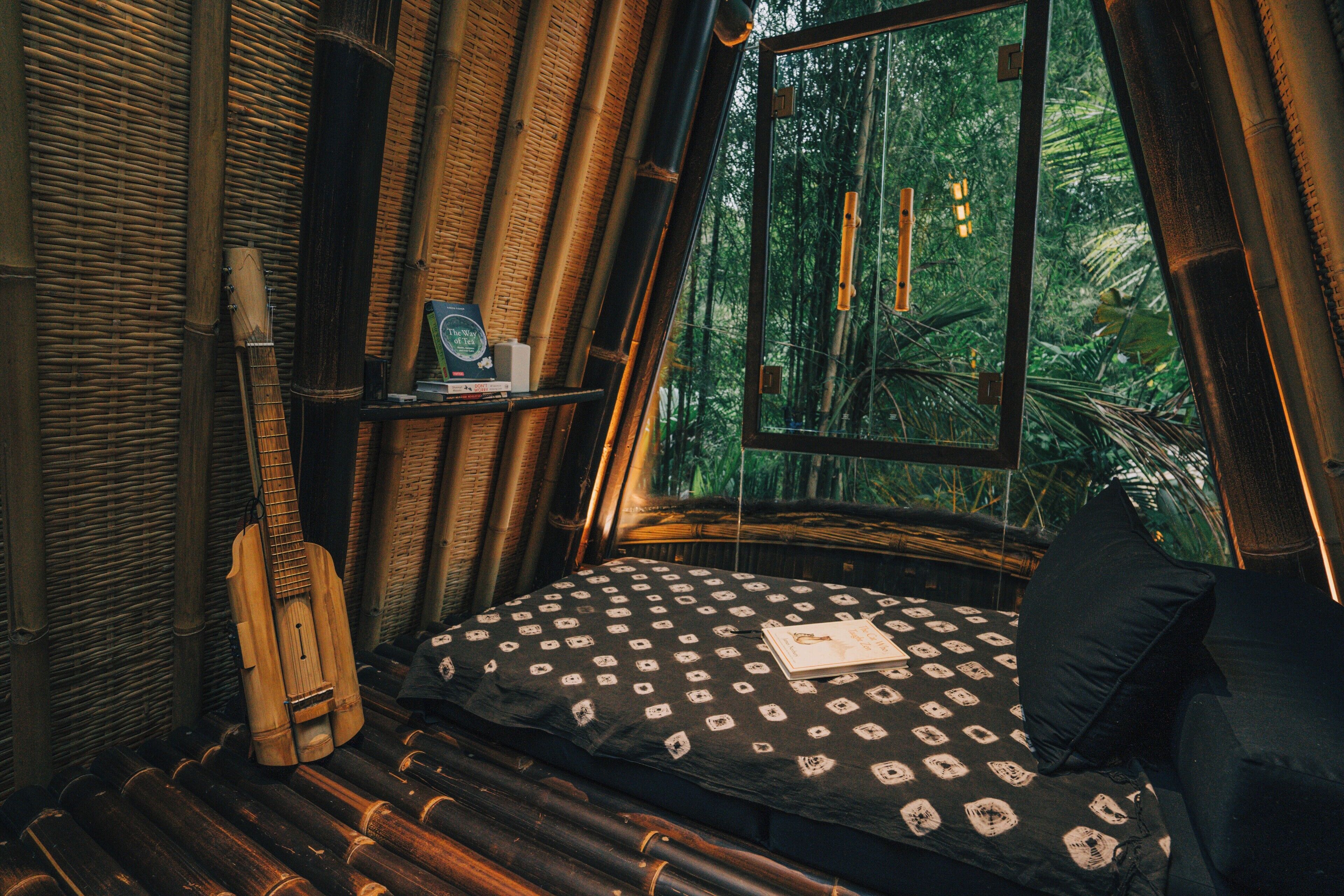 A room with a bed, a guitar, and a window overlooking a forest.