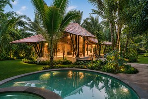 A poolside area with a wooden deck, a pool, and a building with a thatched roof. - Hideout Eco Bamboo House Bali (Selat)