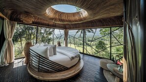 A circular bed with a view of a forested landscape through large windows. - Hideout Eco Bamboo House Bali (Selat)
