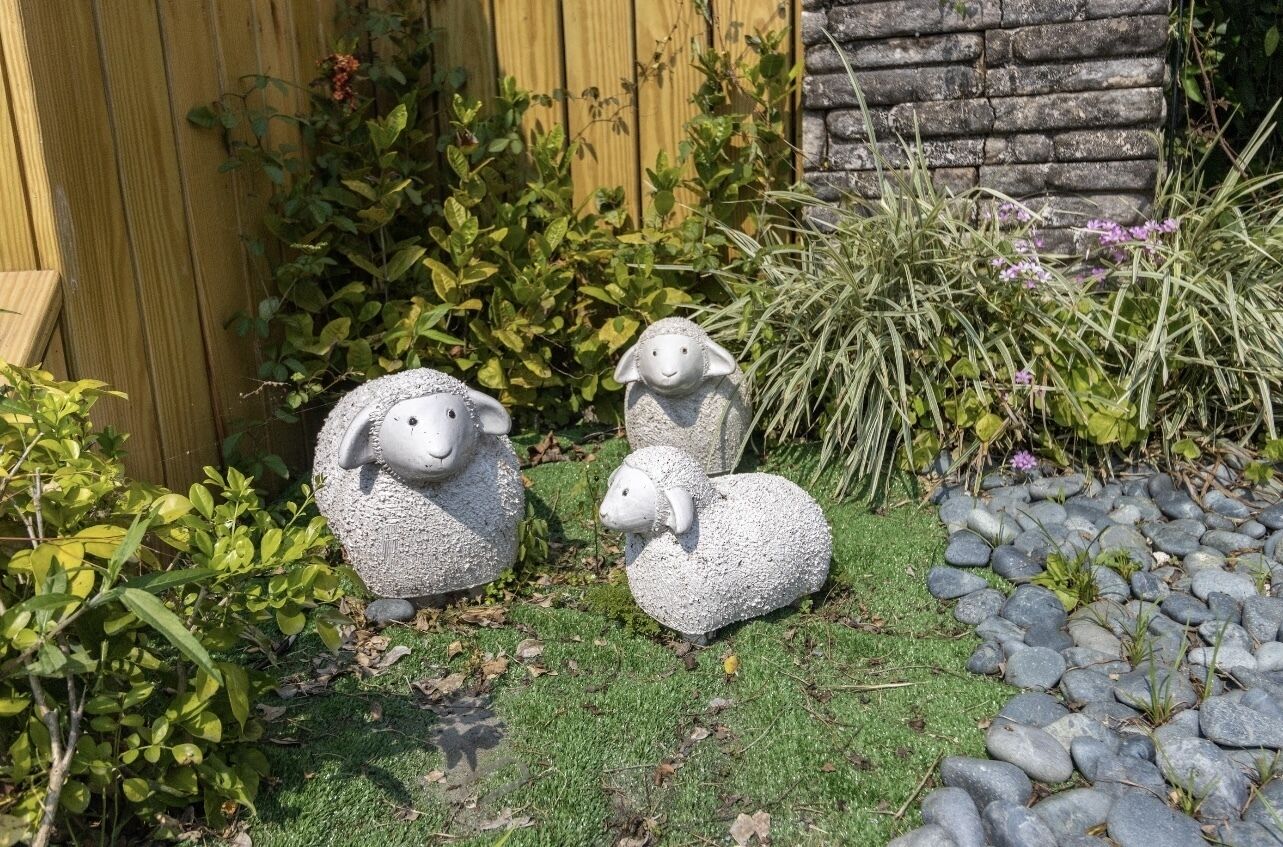 Three stone sheep sculptures in a garden setting with a wooden fence and a stone wall in the background.