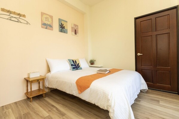 Una cama bien hecha con edredón blanco y manta naranja, una mesita de noche con una caja de pañuelos, y piso de madera.