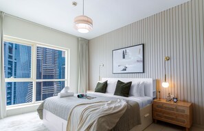 A modern bedroom with a large window, a bed with a white headboard, and a bedside table with a lamp.
