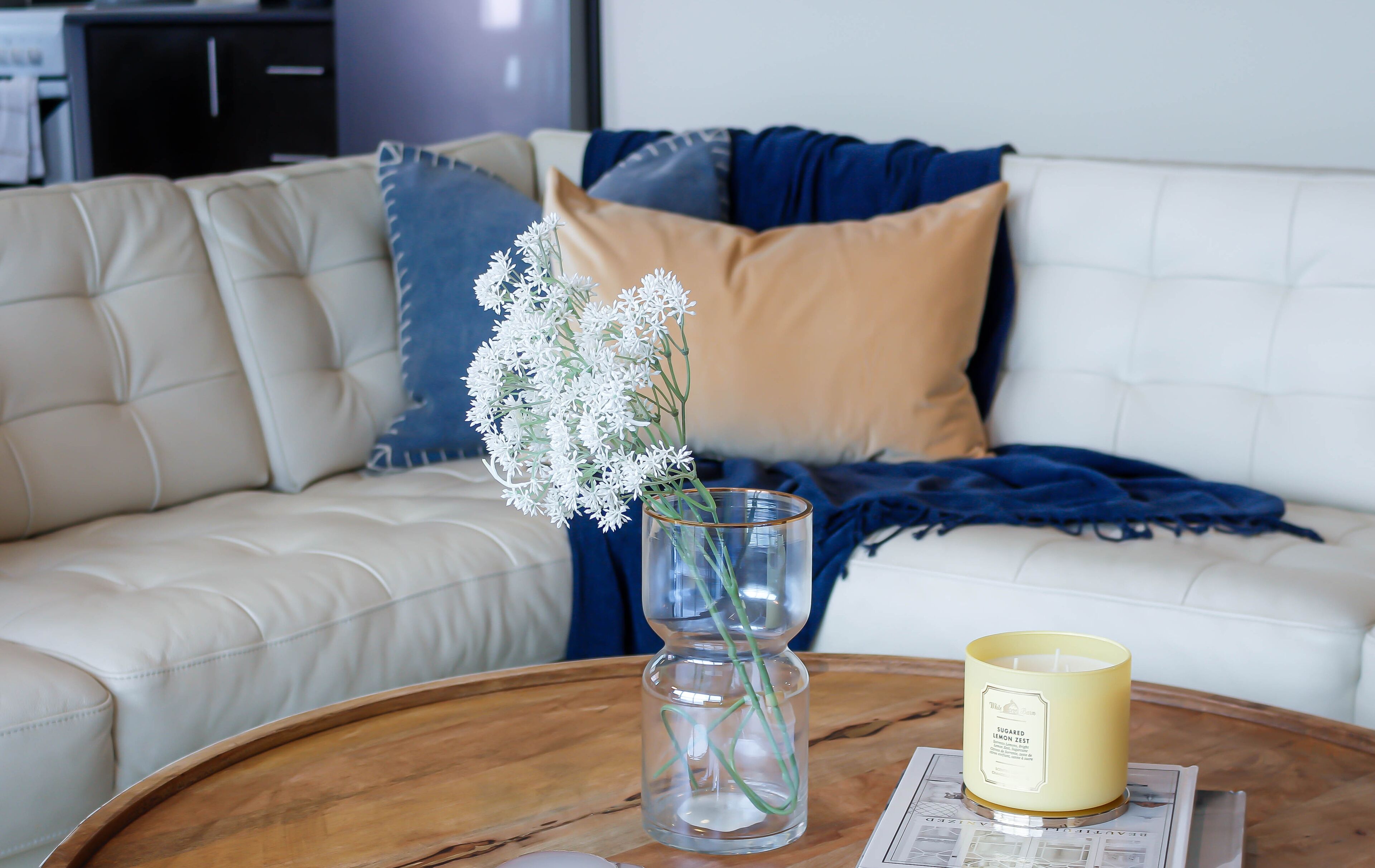 Un salon avec un canapé blanc, un vase en verre contenant des fleurs blanches et une bougie jaune posée sur une table basse en bois.