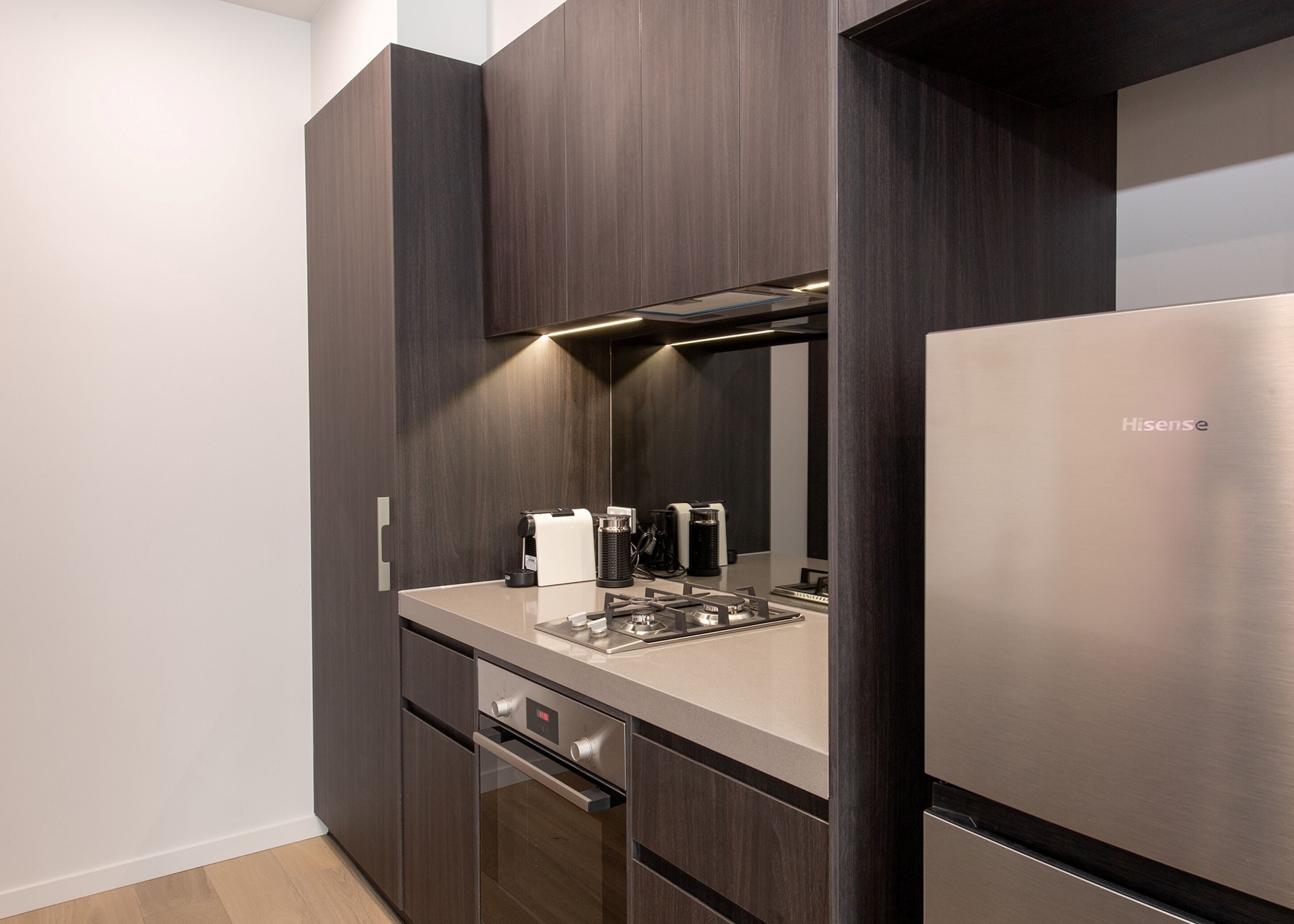 A modern kitchen with dark wood cabinetry, a stainless steel refrigerator, a built-in oven, and a coffee machine.