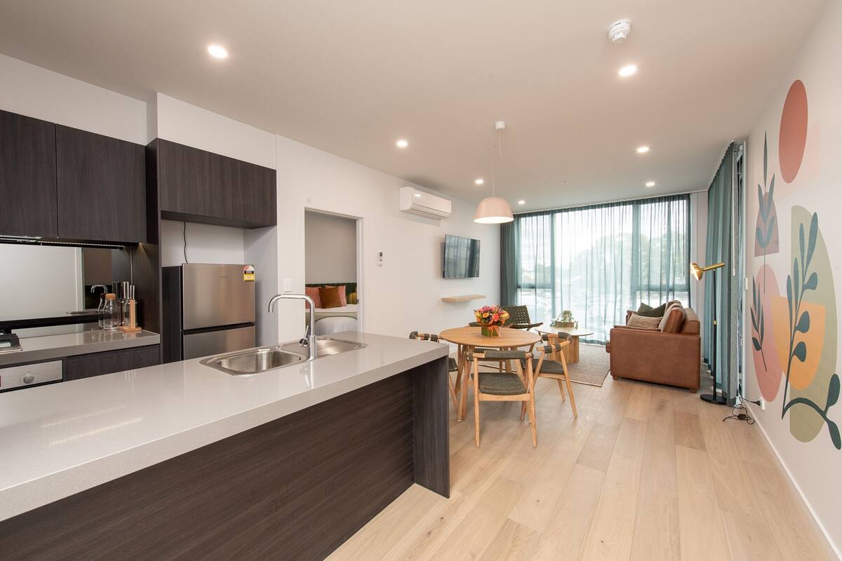 A modern kitchen with dark cabinets, stainless steel appliances, and a central island. There is a dining area with a wooden table and chairs, and a sofa in the living room.