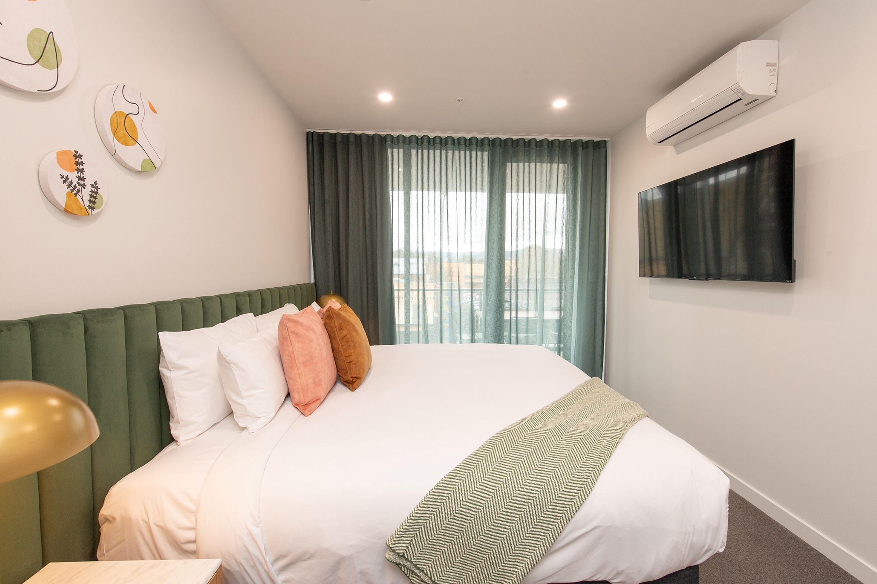 A hotel room with a bed, a flat-screen TV, an air conditioner, and a window with curtains.