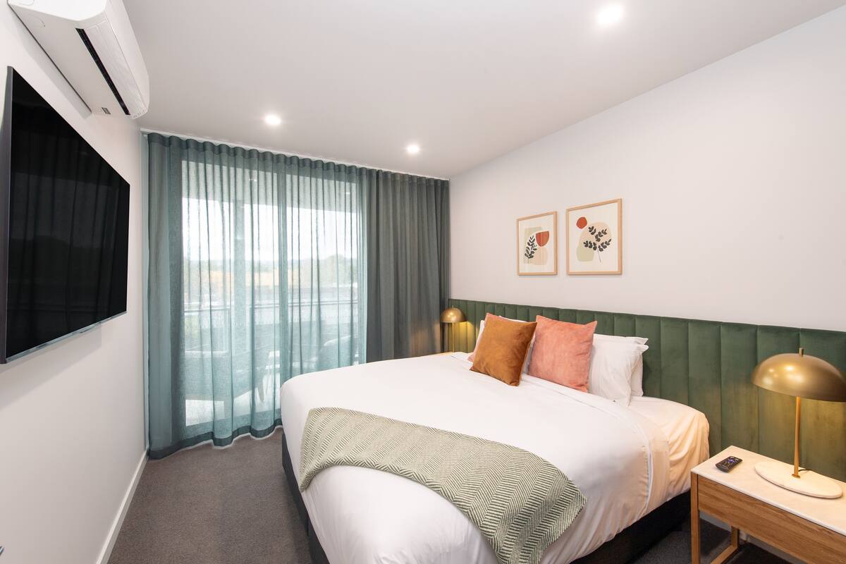 A hotel room with a bed, a nightstand, a lamp, an air conditioner, a TV, and a window with curtains.