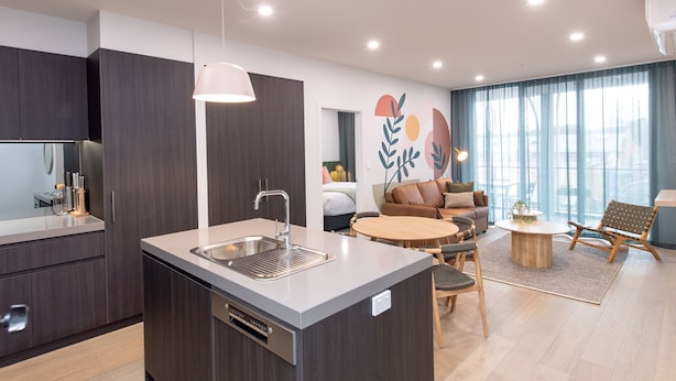 A modern kitchen with a central island, a dining area with a round table and chairs, and a living space with a sofa and a decorative wall. - Grow at Goodwood (Goodwood)