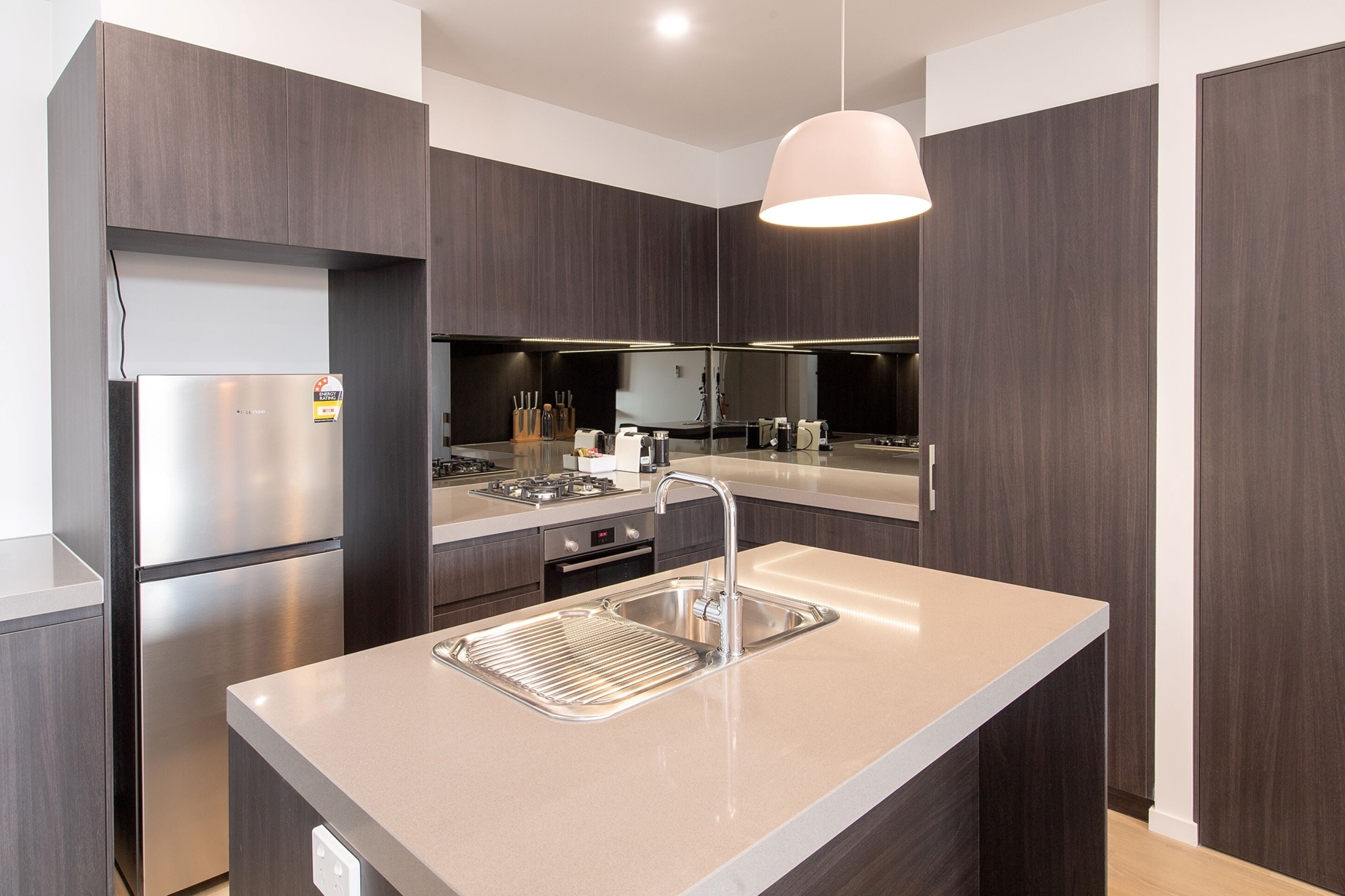 Une cuisine moderne avec des meubles en bois foncé, des appareils en acier inoxydable, un îlot central doté d’un évier et un luminaire suspendu.
