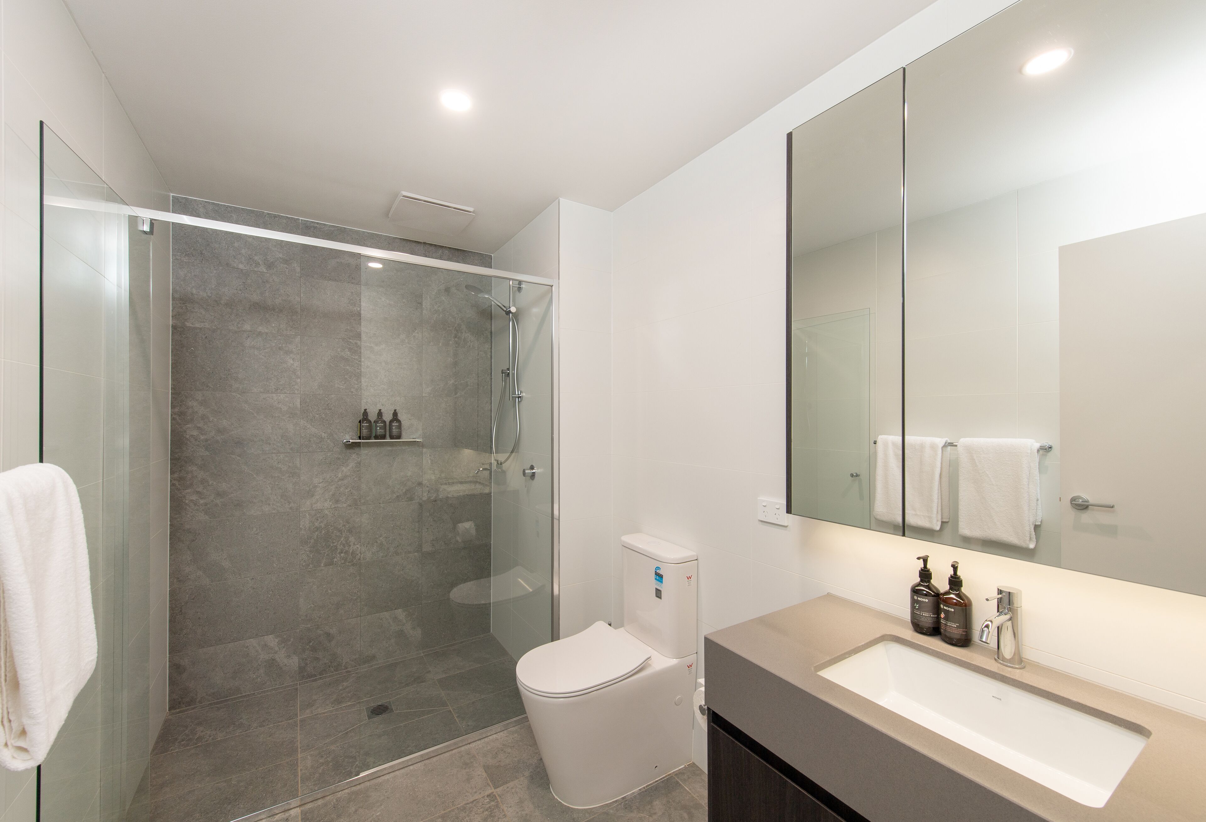 A modern bathroom with a glass-enclosed shower, a white toilet, and a large mirror above a beige sink with toiletries.