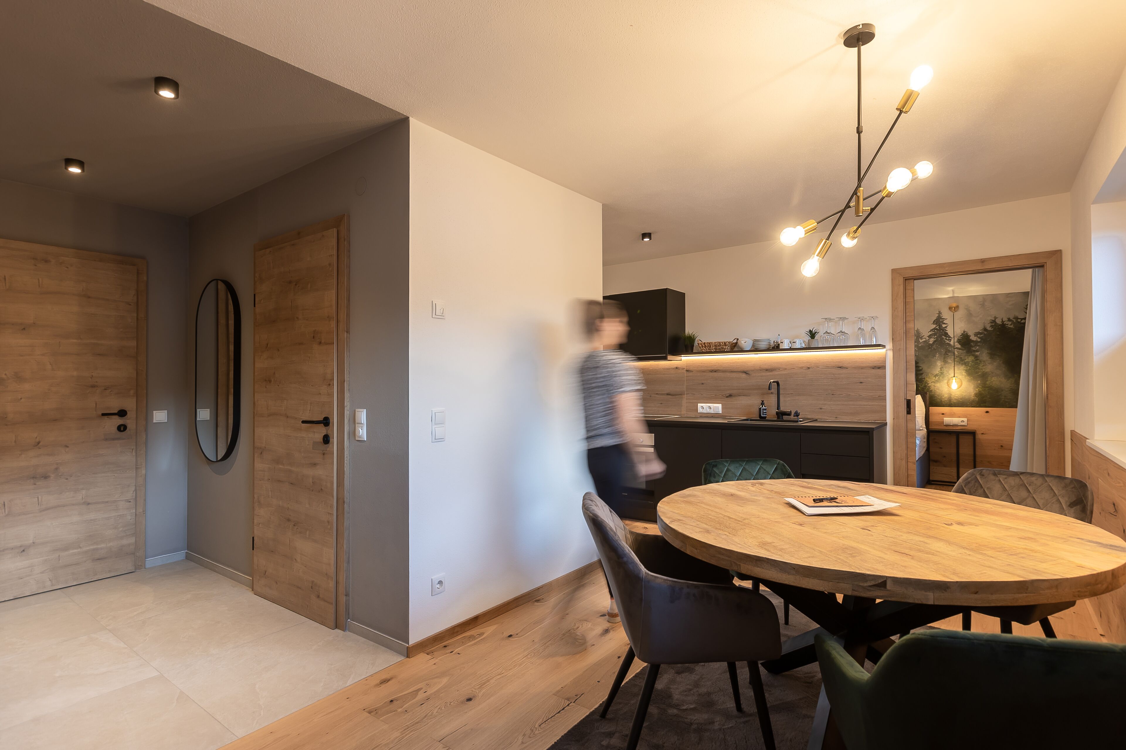 A modern kitchen with a wooden countertop, a dining area with a round wooden table, and a person walking through the space.