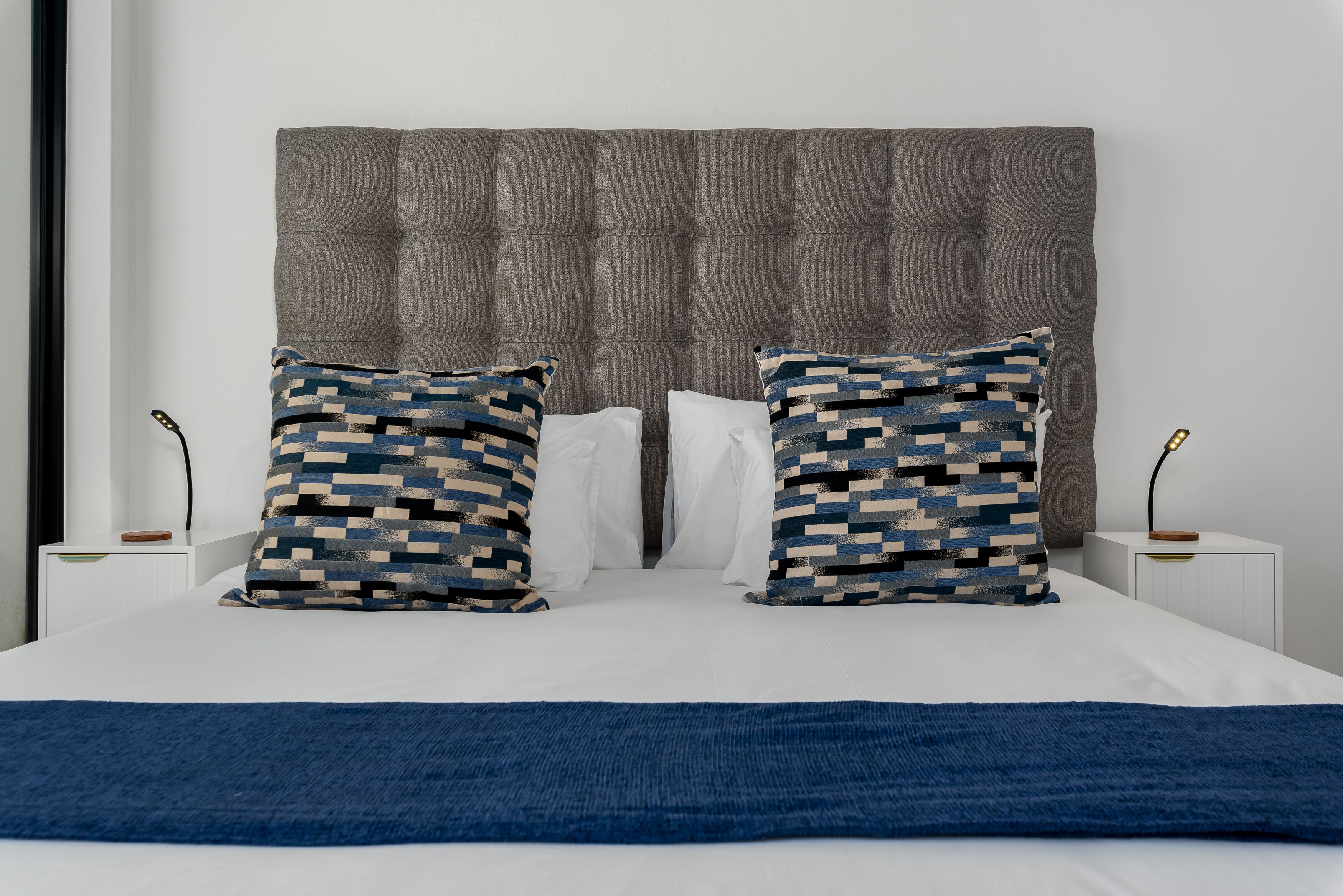 A neatly made bed with a grey upholstered headboard, two patterned pillows, and a blue throw blanket.