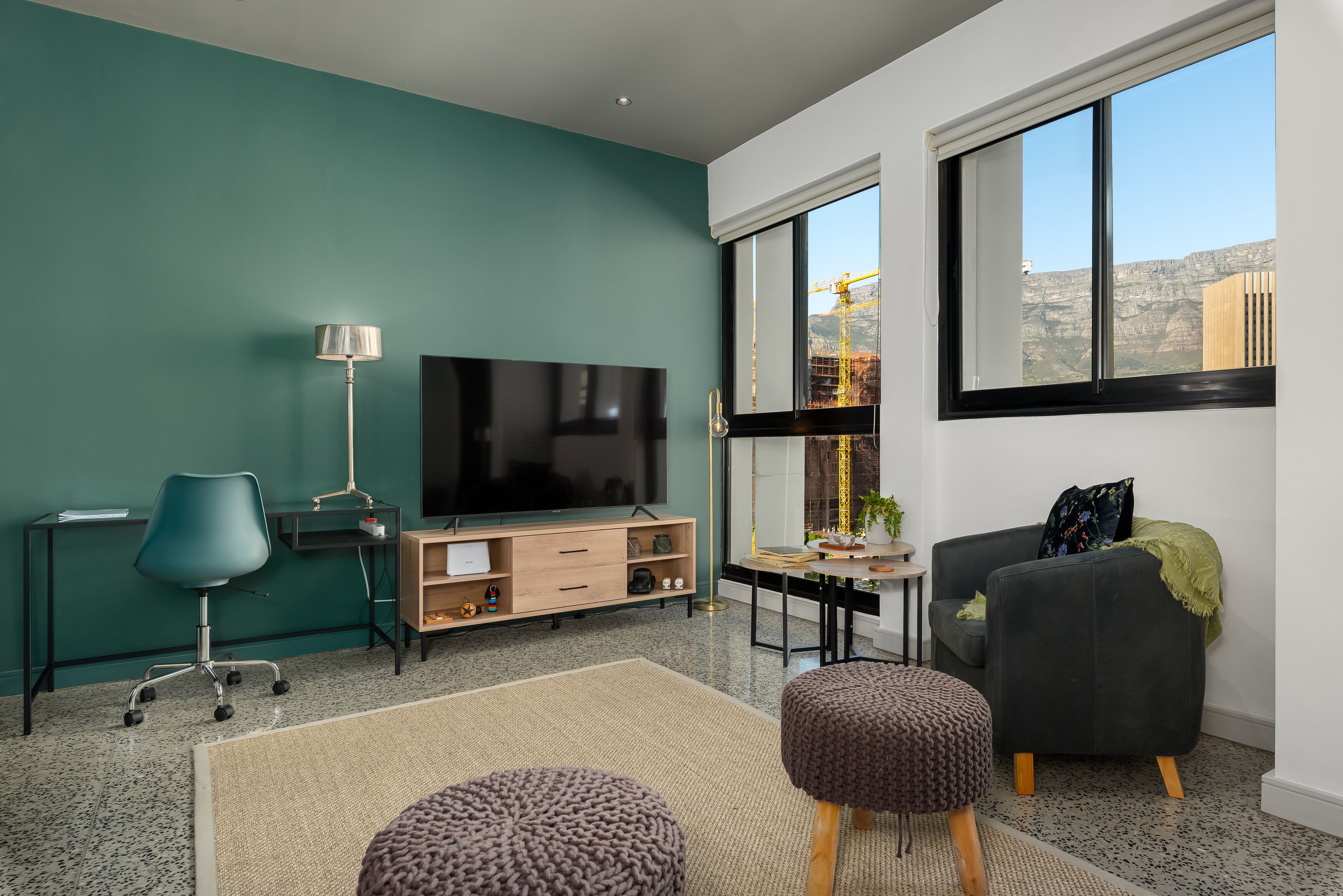 A modern living room with teal walls, a large flat-screen TV, a desk, a comfortable sofa, and a large window with a view of a construction site.