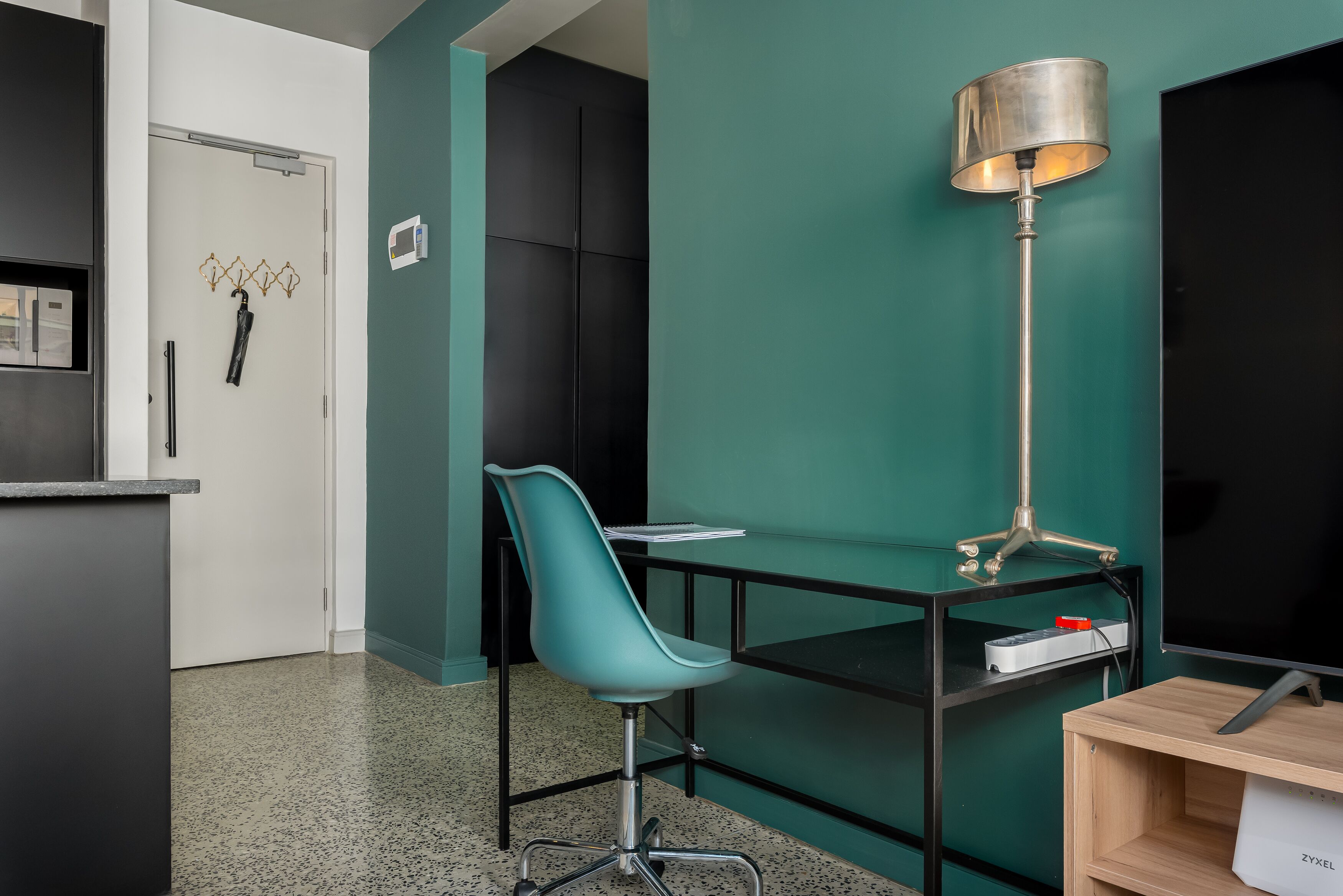 A modern office space with a teal wall, a black desk, a teal office chair, a standing lamp, and a wooden shelf.