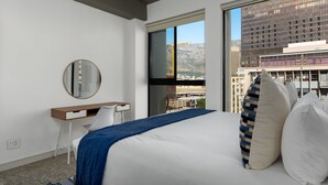 A modern hotel room with a large bed, a desk, a chair, a mirror, and a view of a cityscape through a large window. - The Duke 905 (Cape Town)