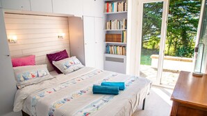 Un dormitorio con cama, almohadas, una estantería y vistas a la vegetación a través de una puerta de cristal.