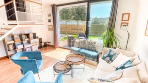 Een ruime woonkamer met een bank, stoelen en een salontafel. Een trap met houten leuning en een boekenplank vol boeken en decoratieve objecten. Een grote schuifdeur leidt naar een buitenruimte met een terrasmeubel en uitzicht op een tuin.