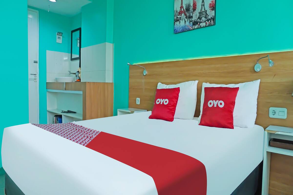 Quarto de hotel com uma cama, mesa de cabeceira, encosto de madeira, colcha vermelha e branca e um quadro da Torre Eiffel.