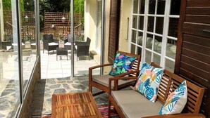 Een zonnig terras met een houten tafel en een bank met kleurrijke kussens, grote ramen en uitzicht op een tuin.