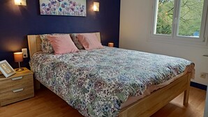 A bedroom with a wooden bed, floral bedding, two wall-mounted lights, a nightstand with a lamp, and a window with a view of trees. - A L'ombre du Frêne, Maison Avec Jardin Arboré. Idéal Pour les Amis de la Nature (Lacombe)