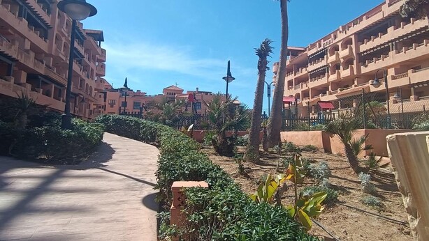 A courtyard with a paved walkway, palm trees, and buildings with balconies. - Apartment in Almerimar with Pool & Beaches (Almeria)