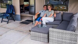 A couple sitting on a grey sofa in a covered outdoor area with wicker furniture and a glass table.