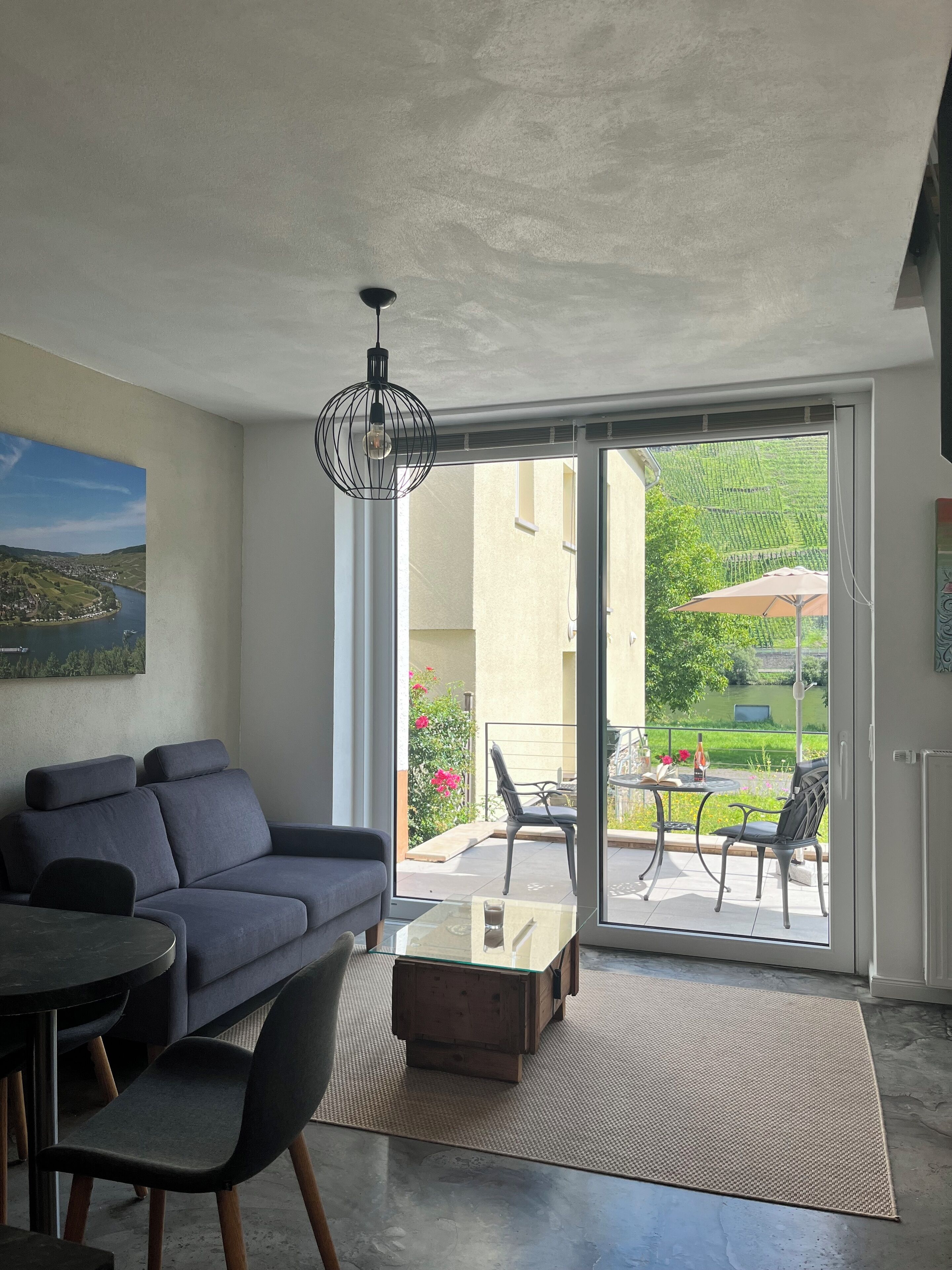 A modern living room with a sofa, coffee table, and chairs, a dining table, and a large sliding glass door leading to an outdoor patio with a garden view.
