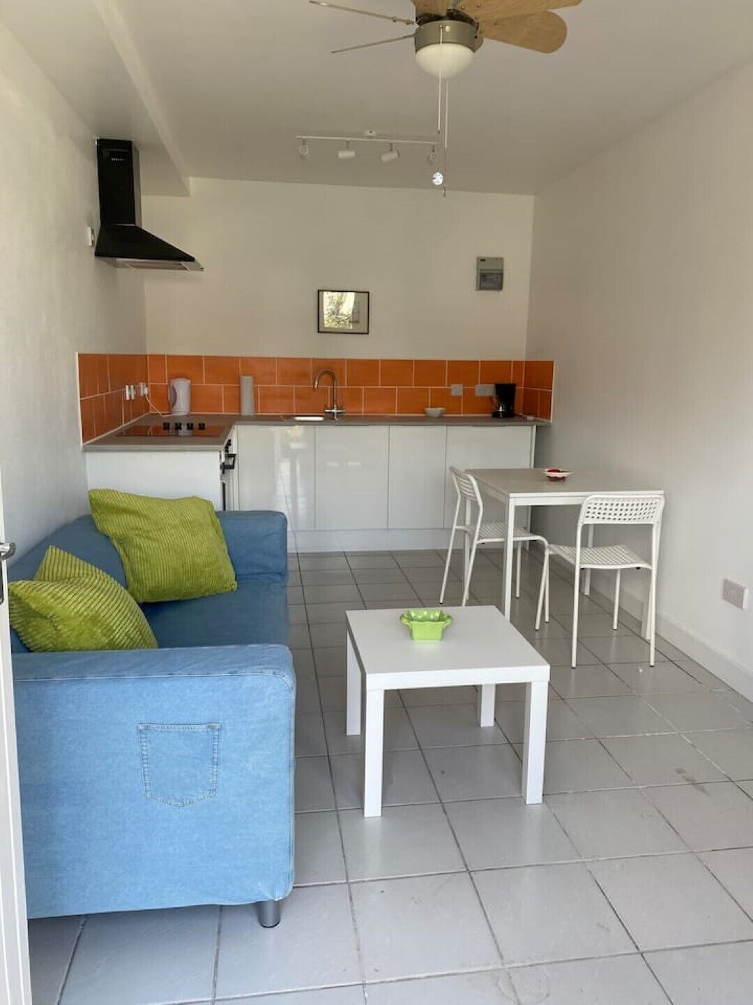 A compact living space with a kitchenette, a blue sofa with green cushions, a white table with a green bowl, and a ceiling fan.