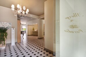 En hotellkorridor med ett svart- och vitrutigt golv, en kristallkrona, en vägg med guldfärgad dekorativ skrift och en krukväxt.