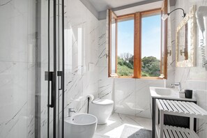 A modern bathroom with a glass shower enclosure, a toilet, a sink, and a window with a view of greenery.
