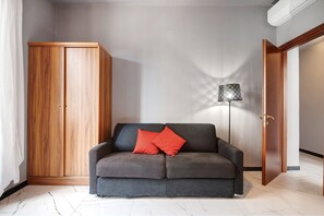 Ett rum med en grå soffa, en trägarderob, en golvlampa och en dörr.