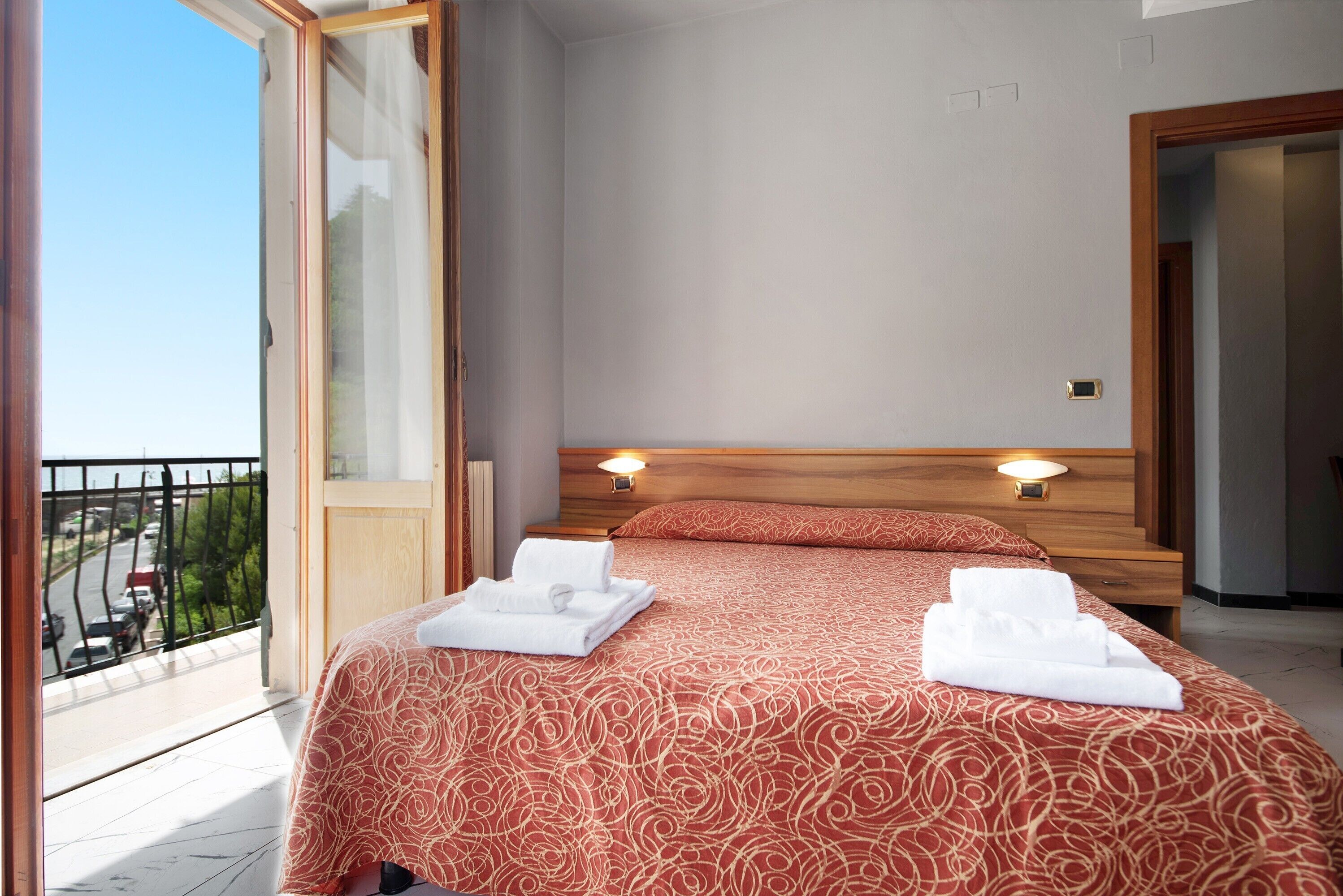 Habitación de hotel con balcón, cama con colcha estampada, dos lámparas de noche y cabecero de madera.