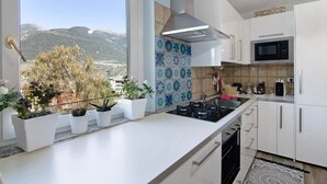 Uma cozinha moderna com armários brancos, micro-ondas de aço inoxidável e vista para montanhas através de uma grande janela.