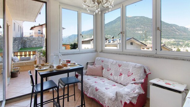 Quarto com mesa de jantar, cadeiras, sofá, lustre e amplas janelas com vista para as montanhas.