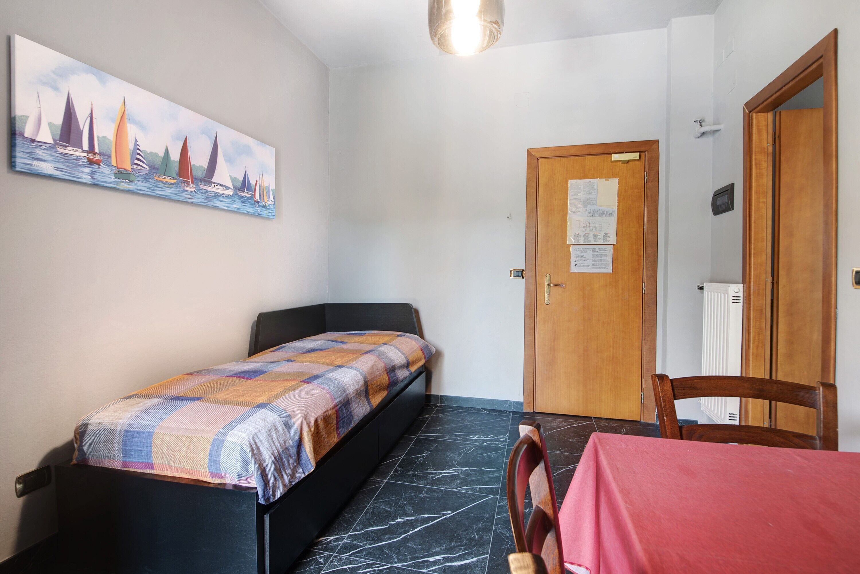 Une chambre à coucher avec un lit, un tableau représentant des voiliers, une porte en bois et une table recouverte d’une nappe rouge.