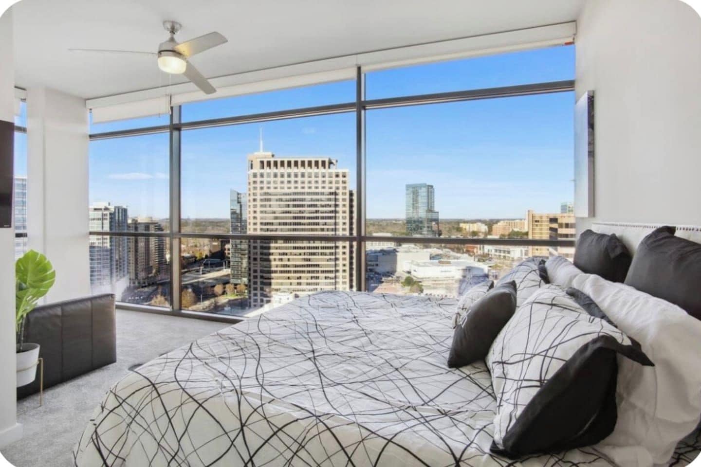 Een slaapkamer met een groot bed, een plafondventilator en een uitzicht op een stadsbeeld door grote ramen.