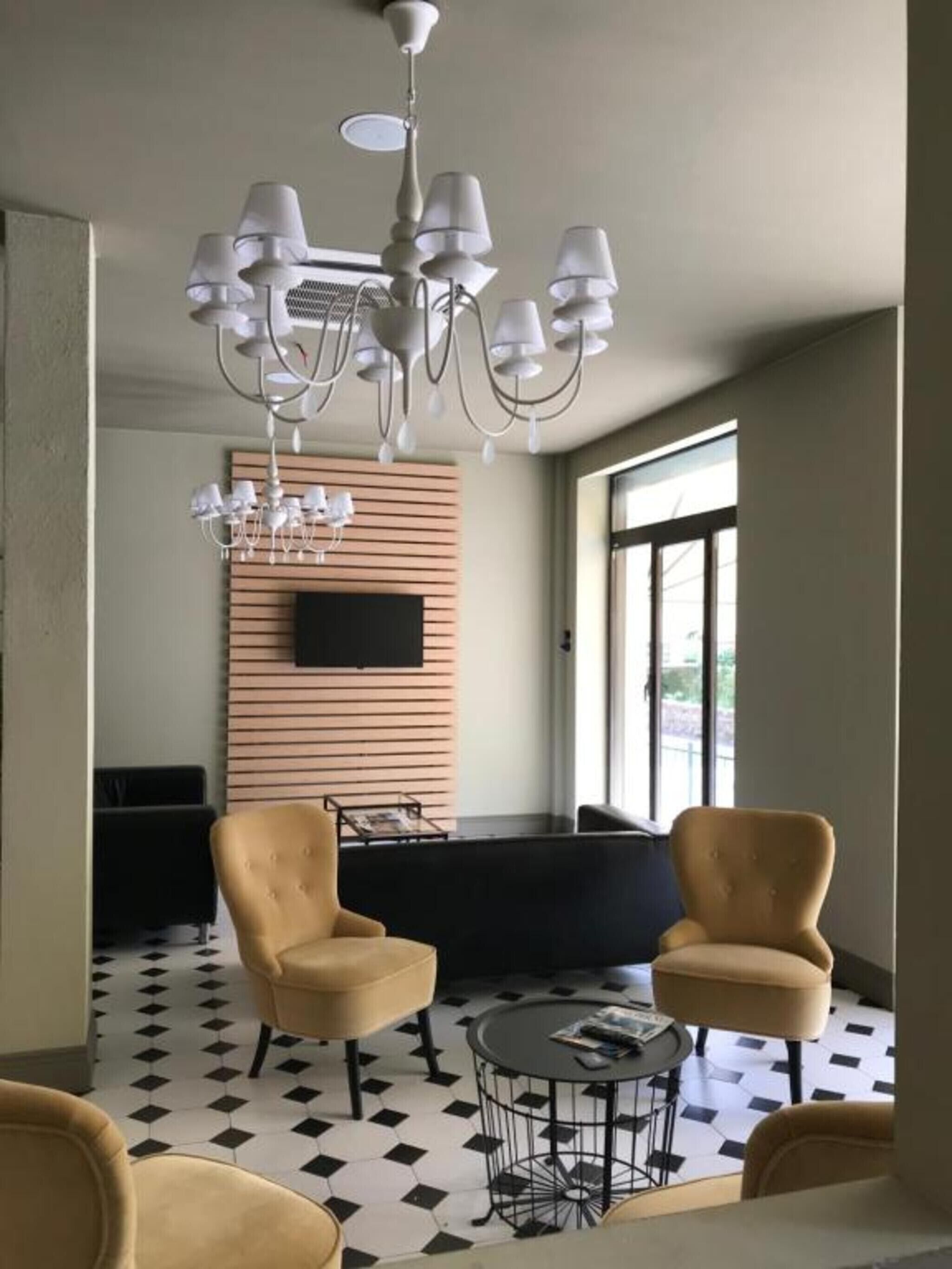 Een moderne woonkamer met een kroonluchter, twee fauteuils, een ronde salontafel, een flatscreen televisie bevestigd aan een houten paneel, en een groot raam.